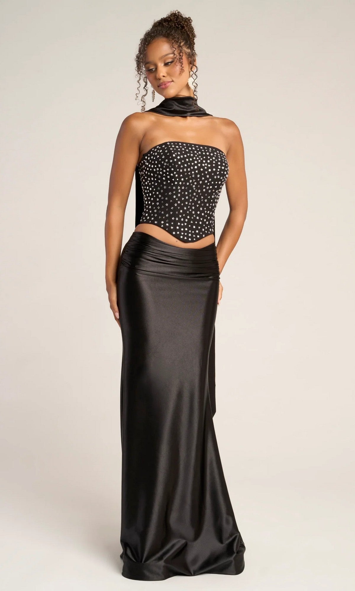 A woman stands against a plain background wearing the Portia and Scarlett PS26042, a long formal dress featuring a strapless black beaded corset top with choker-style scarf and fitted satin skirt.